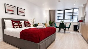 Habitación de hotel moderna con una cama grande, un área de comedor con mesa y sillas, y vista a la ciudad desde la ventana.