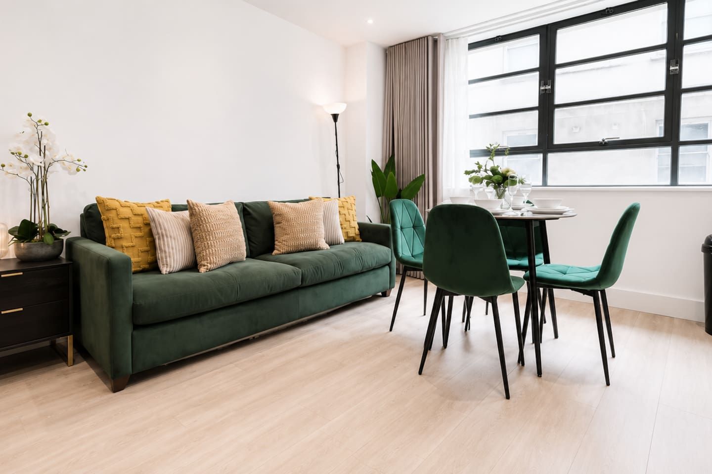 A modern living room with a green sofa, matching chairs, a dining table, and a large window with curtains.