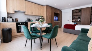 Cozinha moderna com armários de madeira, área de jantar com cadeiras verdes, televisão e cama com cobertor vermelho.