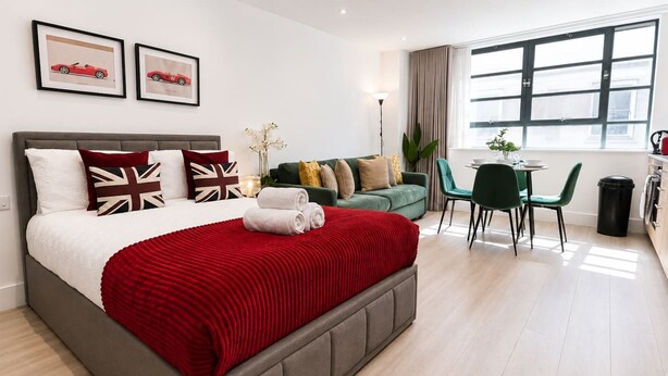 Una habitación de hotel con cama, sofá verde, mesa de comedor con sillas y ventana con cortinas.