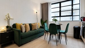 Ein modernes Wohnzimmer mit einem grünen Sofa, einem Esstisch mit Stühlen, einer Stehlampe und einem großen Fenster mit Vorhängen.