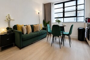Una sala de estar moderna con un sofá verde, una mesa de comedor con sillas, una lámpara de pie y un ventanal con cortinas.
