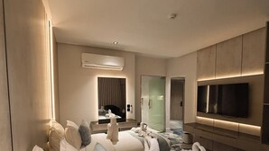 Una camera d'albergo moderna con un letto grande, una TV a schermo piatto, un condizionatore d'aria e un bagno visibile tramite una porta in vetro.