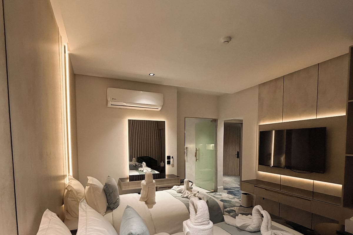 A modern hotel room with a large bed, a flat-screen TV, an air conditioning unit, and a bathroom visible through a glass door.