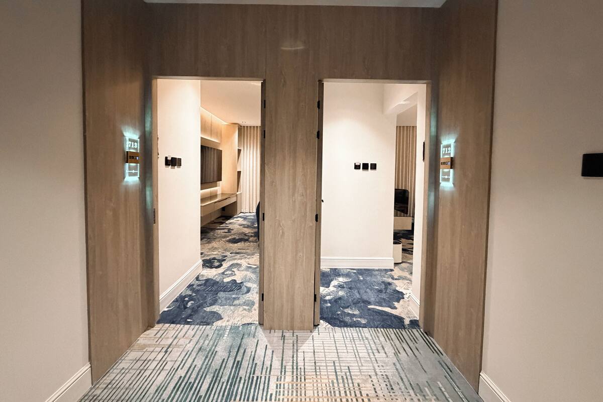 A hotel hallway with wooden doors, a patterned carpet, and a bed visible in the adjacent room.