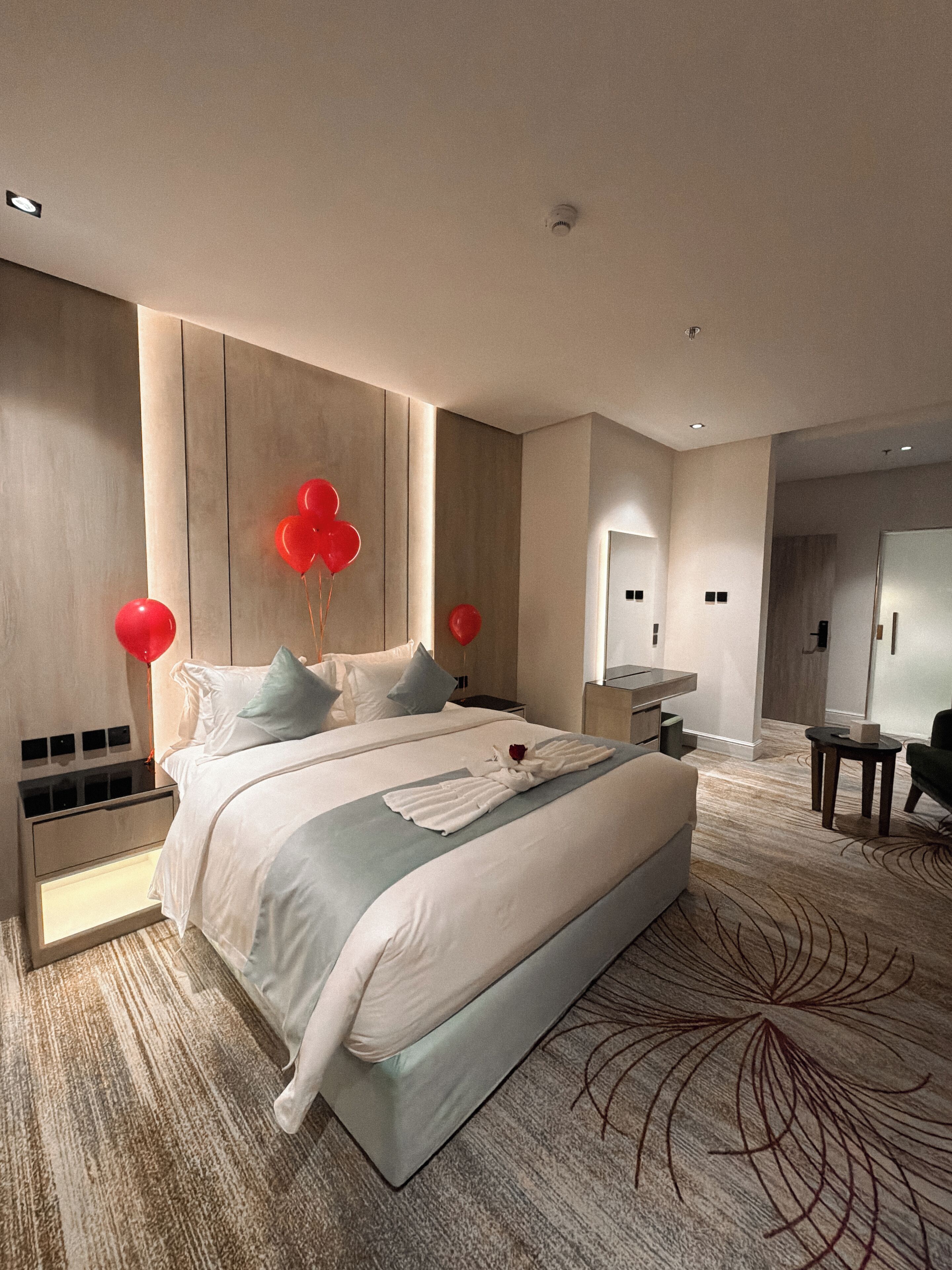 A modern hotel room with a large bed, a bedside table, a nightstand, a mirror, and a small seating area.