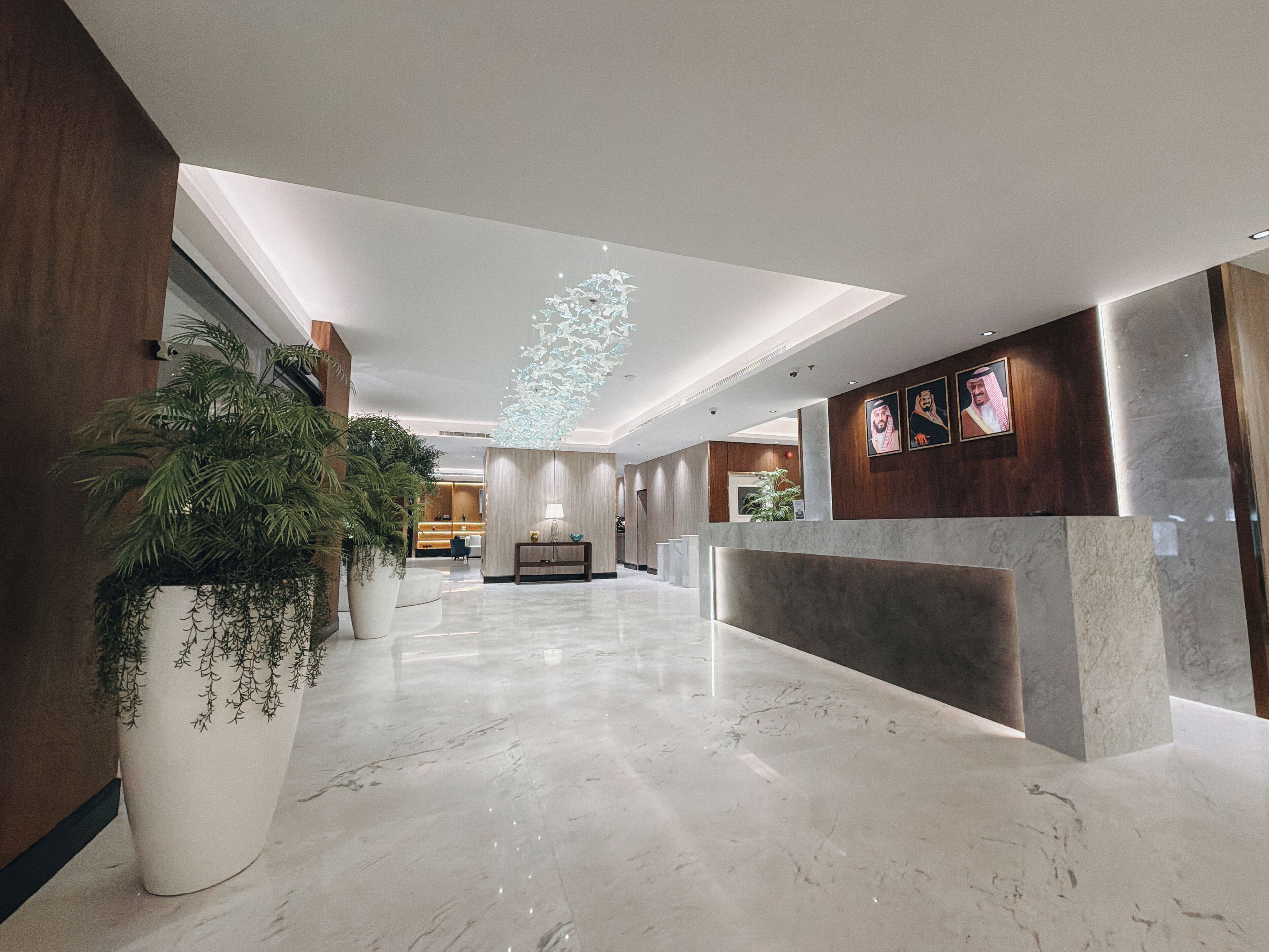 Un vestíbul d'hotel modern amb sòl de marbre, una taula de recepció, una gran planta en test i una làmpada de sostre decorativa.