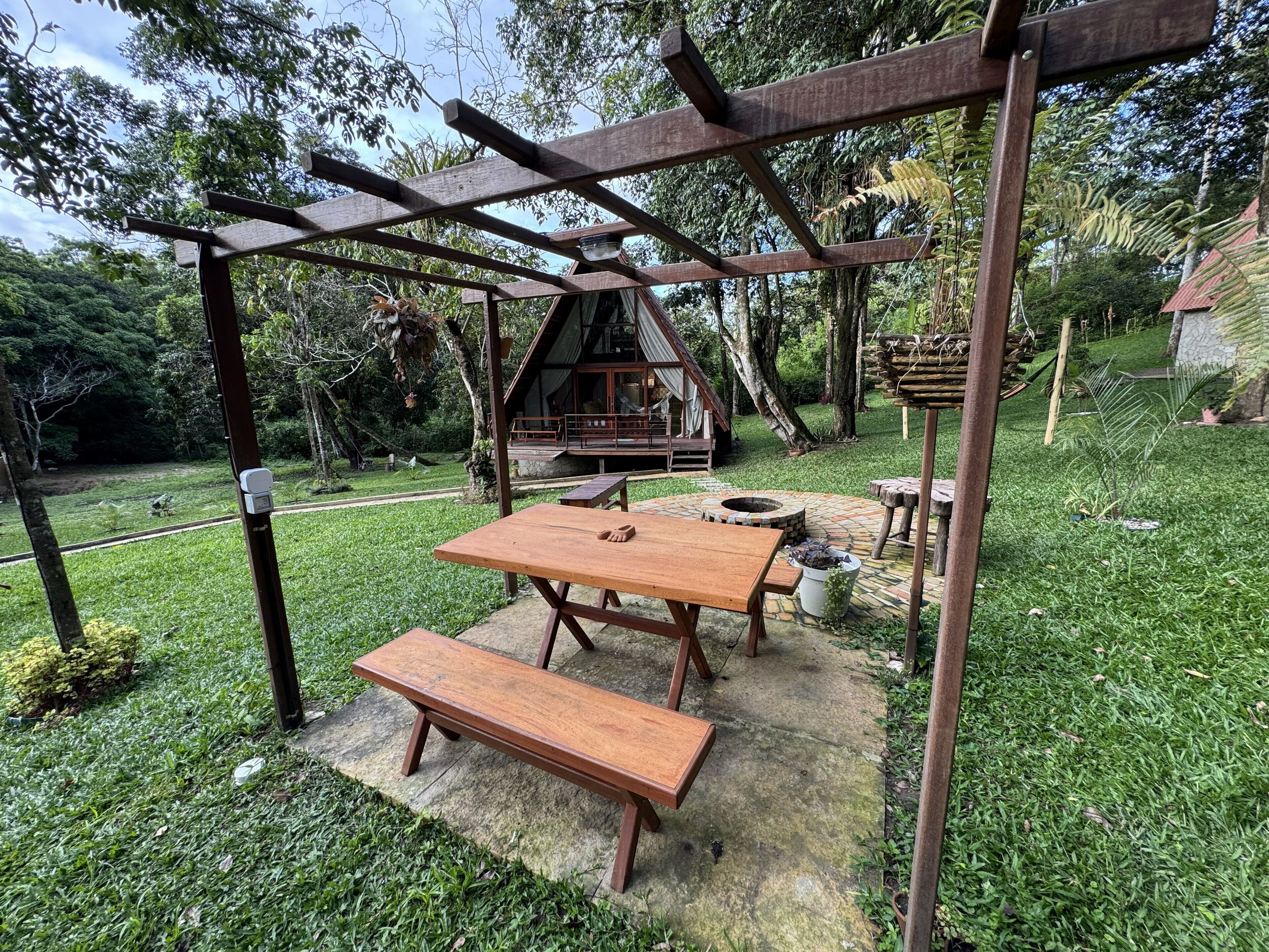 Una pérgola de madera con una mesa y banco, un hogar a fuego de piedra, y una cabaña rústica al fondo.