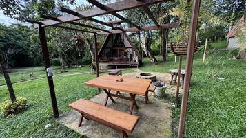 Una pérgola de madera con una mesa y banco, un hogar a fuego de piedra, y una cabaña rústica al fondo.