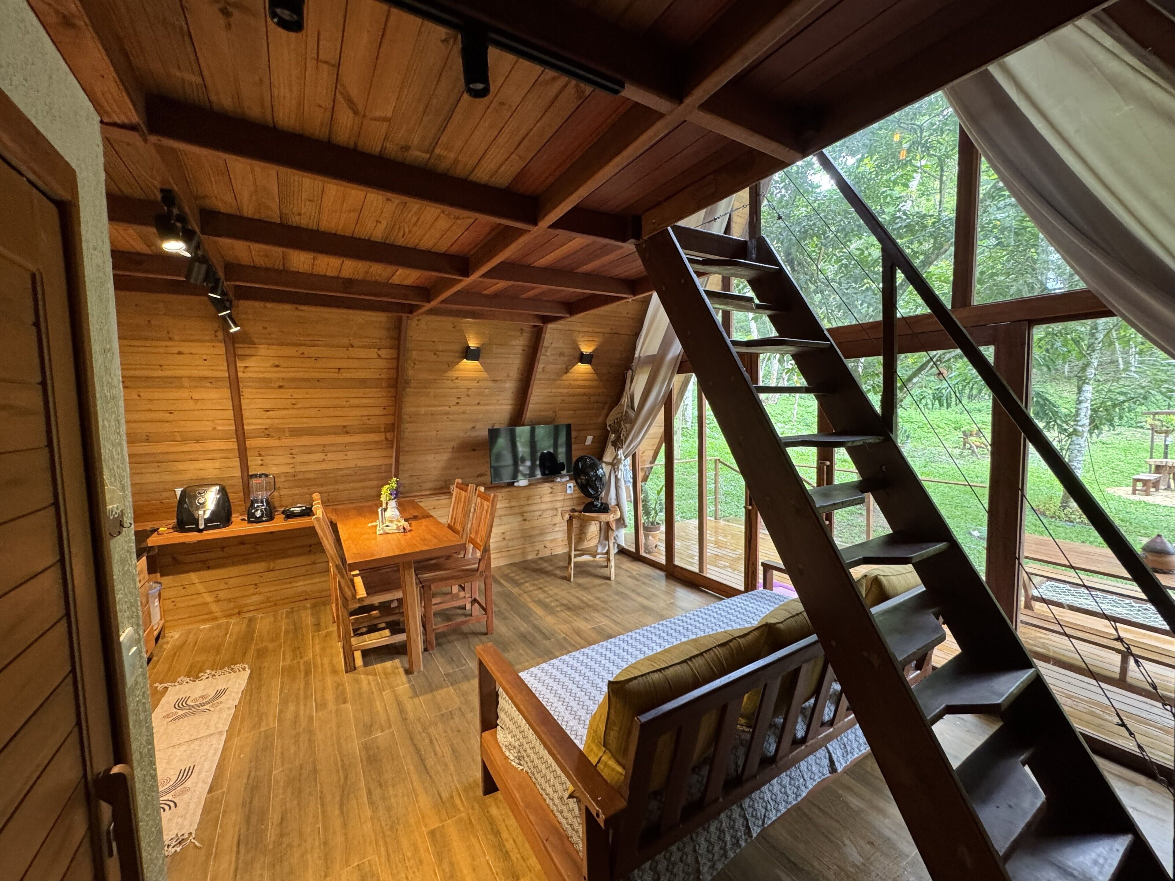 A wooden interior with a dining area, a bed, a TV, and a staircase leading to a balcony.