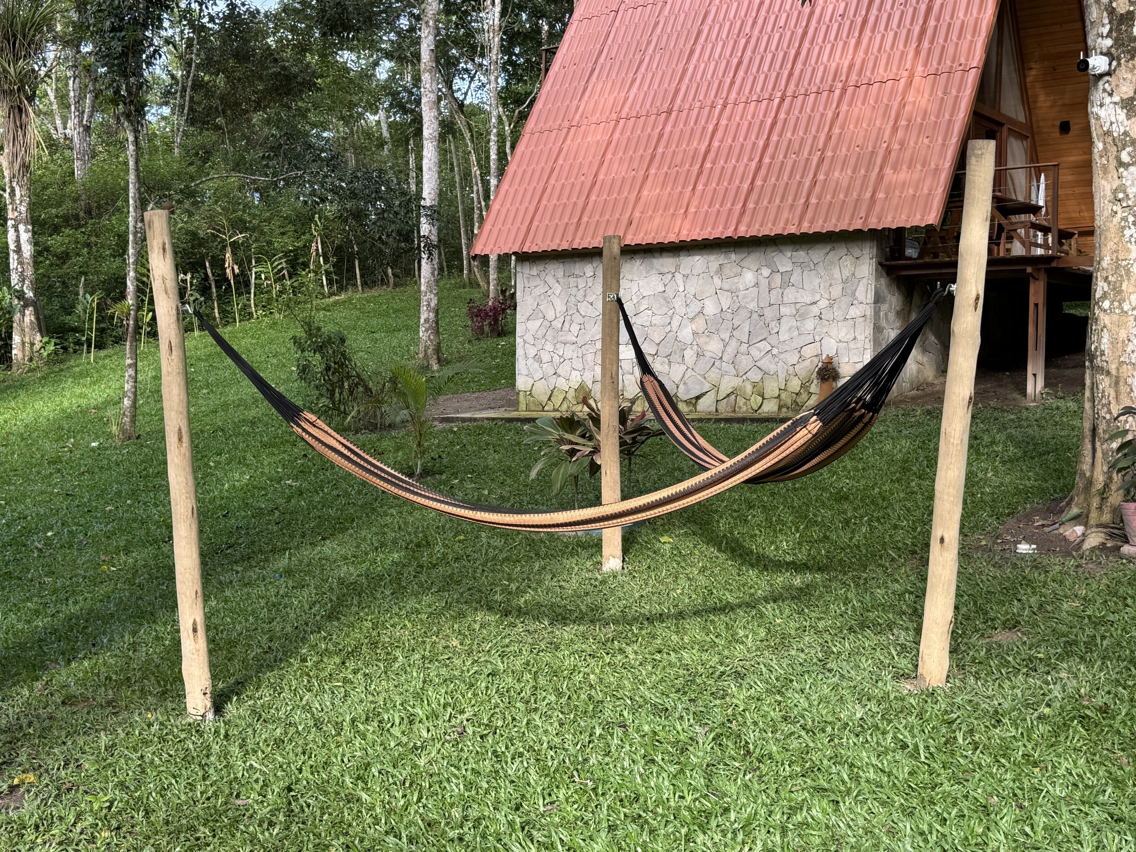 A hammock between two wooden poles in a grassy area with a stone building and a red roof in the background.