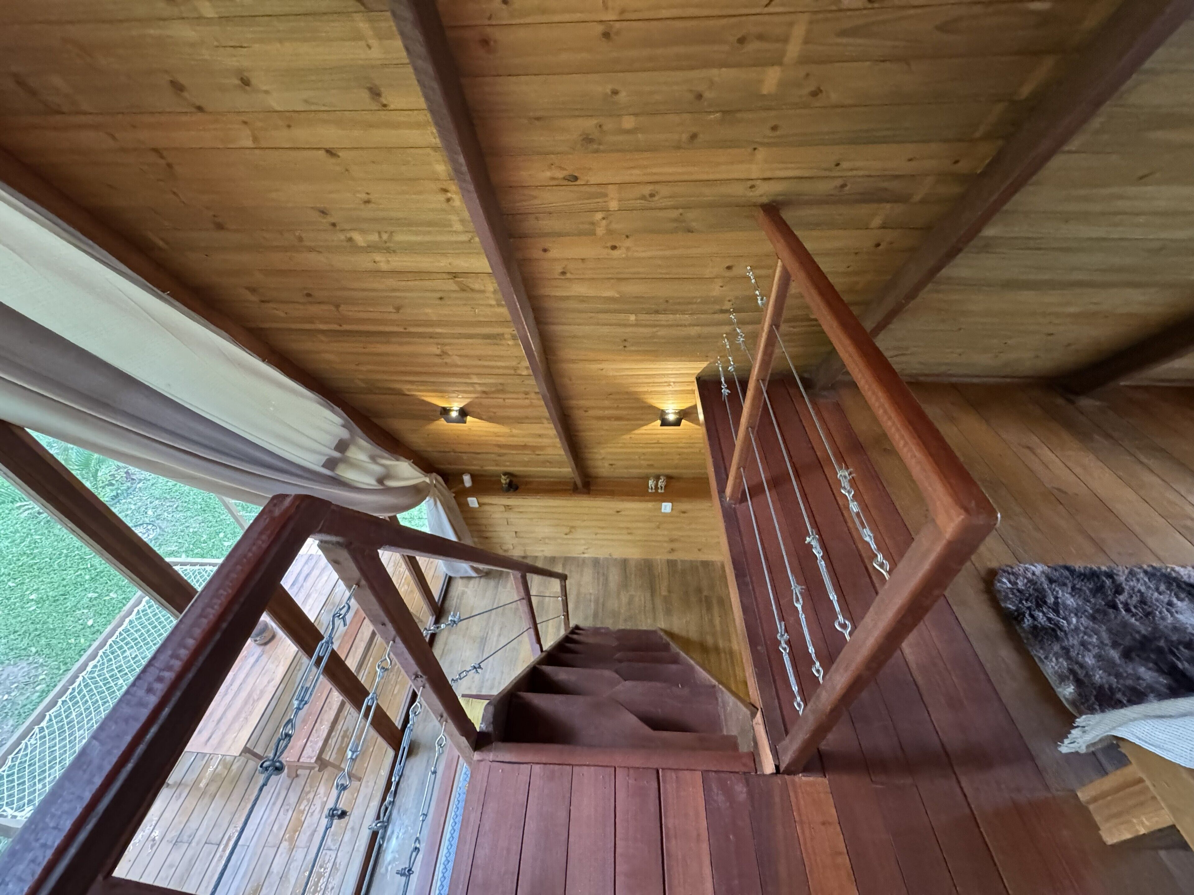 A wooden staircase with a metal railing, wooden beams on the ceiling, and a view of greenery outside.