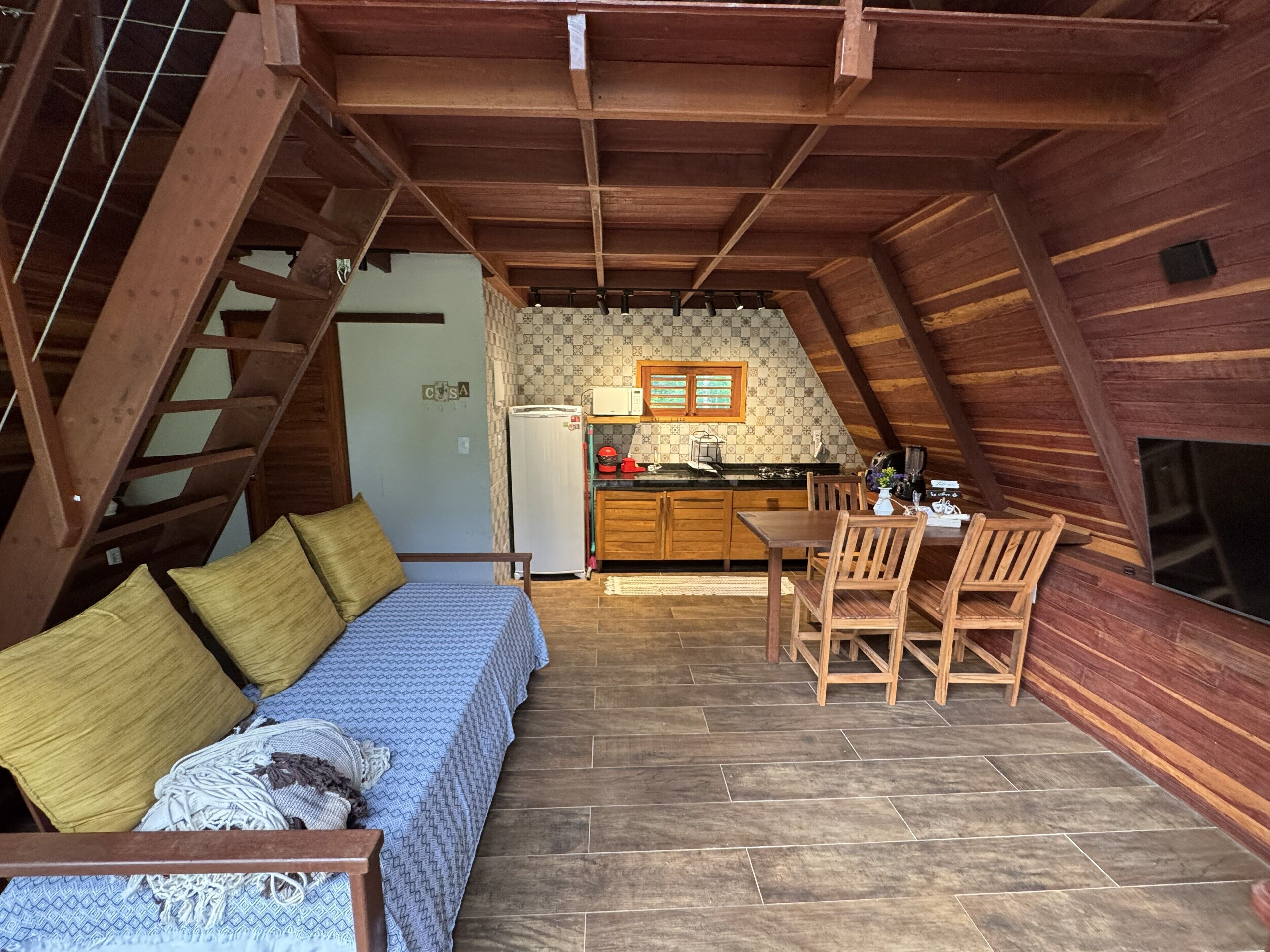 A loft-style bedroom with a bed, a wooden staircase, and a kitchenette with a refrigerator and stove.