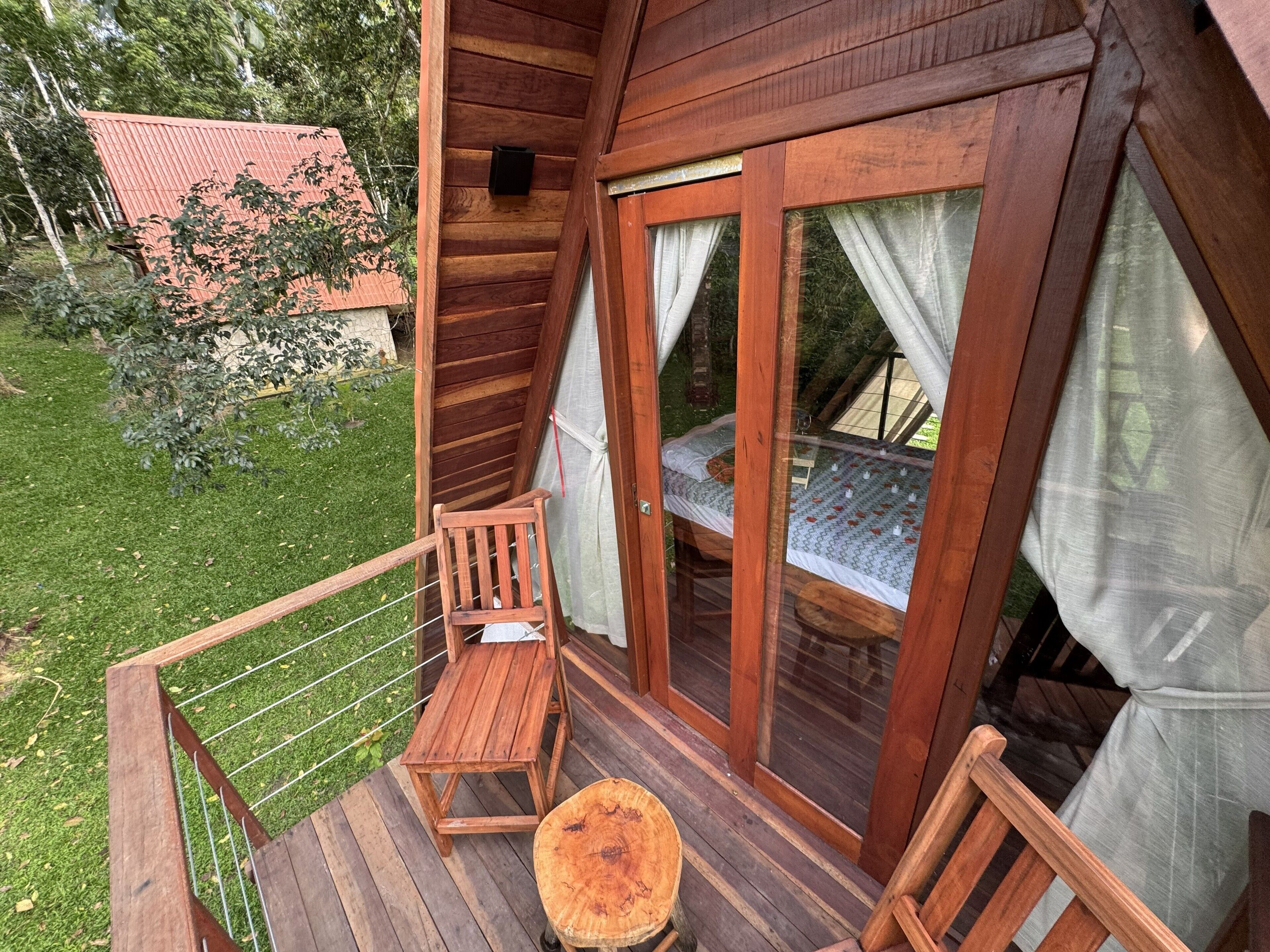 A wooden cabin with a balcony, a wooden chair, a small table, and a bed with a patterned bedspread.