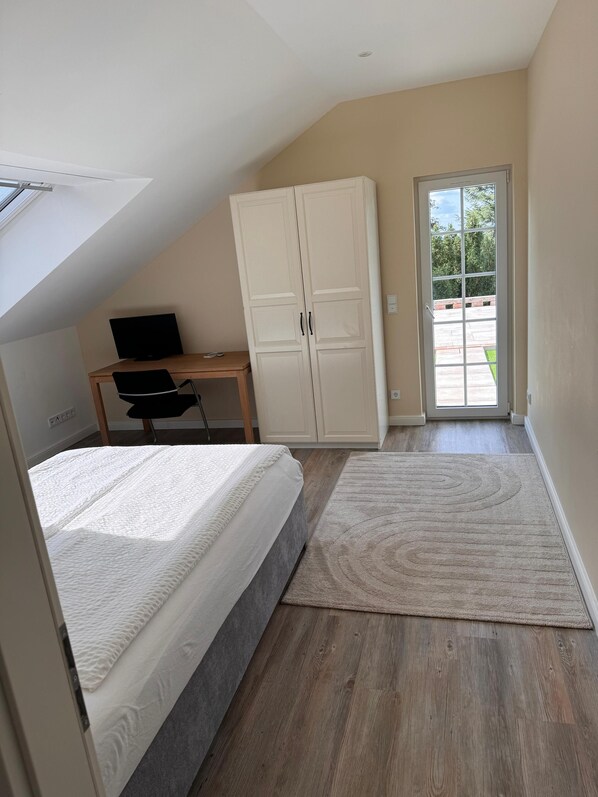 A bedroom with a bed, a desk with a computer, a wardrobe, and a view of the outdoors through a door.