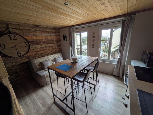 Ein gemütliches Zimmer mit Holzbalkendecke, einer großen Wanduhr, einem Holztisch mit Stühlen, einer Couch und einem Fenster mit Vorhängen.