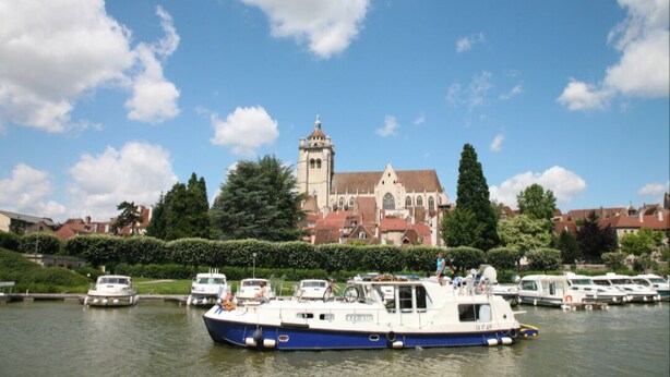 A river with boats, a church with a tower, and residential buildings in the background. - La Darbella (Dole)