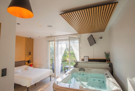 A modern hotel room with a bed, a large window, a sliding glass door leading to a balcony, and a hot tub.