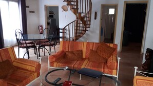 Un salon comprenant un canapé, des fauteuils, une table basse en verre, un escalier en colimaçon et un coin repas avec des chaises et une table.