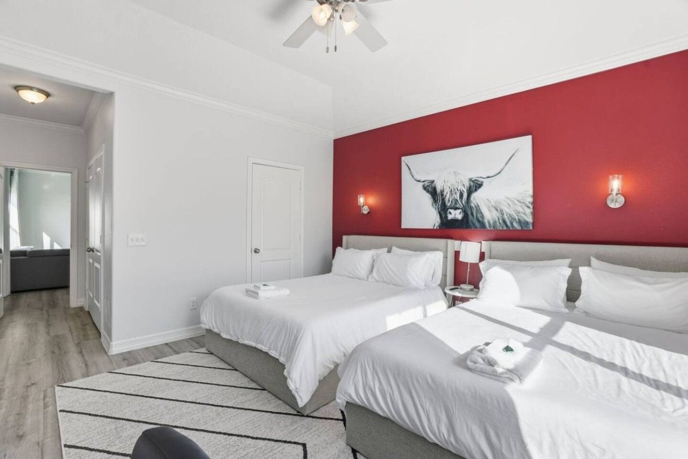 A hotel room with two beds, a red accent wall, a ceiling fan, and a large window.