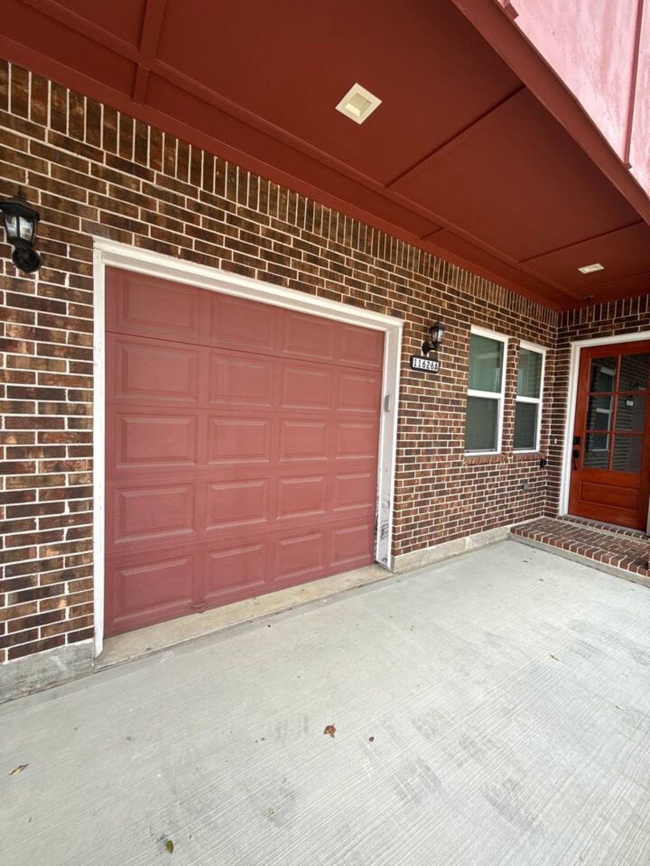A residential garage with a red door, a brick wall, and a small porch area.