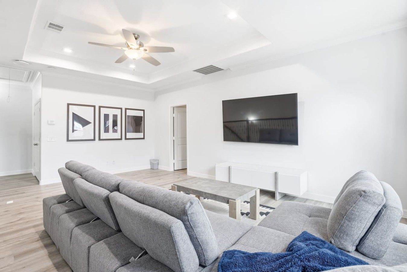 A modern living room with a grey sectional sofa, a flat-screen TV mounted on the wall, and a ceiling fan.