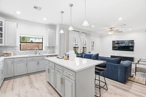 A modern kitchen with white cabinetry, a central island, pendant lighting, and a dining area with blue upholstered furniture. - SW Houston 3BR | Sleeps 10, Yard + 2-Car Garage (Houston)