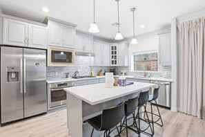 Una cocina moderna con armarios blancos, electrodomésticos de acero inoxidable, una isla central con taburetes de bar, iluminación colgante y una ventana con vista a un edificio de ladrillo.