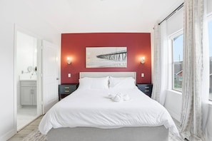 A bedroom with a bed, two bedside tables, a nightstand, a window with curtains, and a painting on the wall.