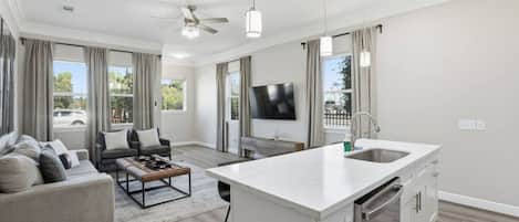 A modern living room with a sofa, coffee table, and TV. There's a kitchen with a large island and a dining area.
