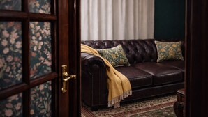 Un sofá de cuero marrón oscuro con una manta amarilla y almohadas estampadas, situado frente a una cortina blanca y una puerta con papel tapiz floral.