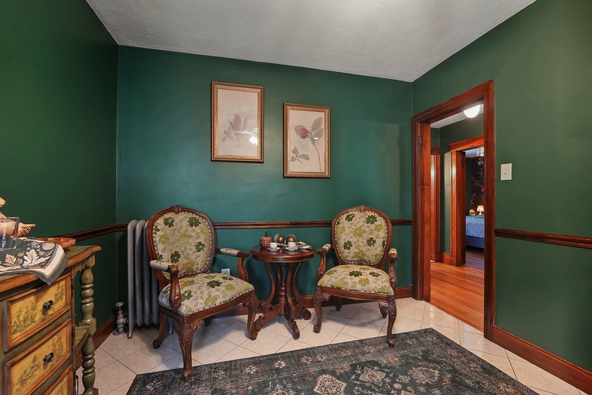 A room with teal walls, two framed floral paintings, a wooden table with chairs, and a vintage dresser.