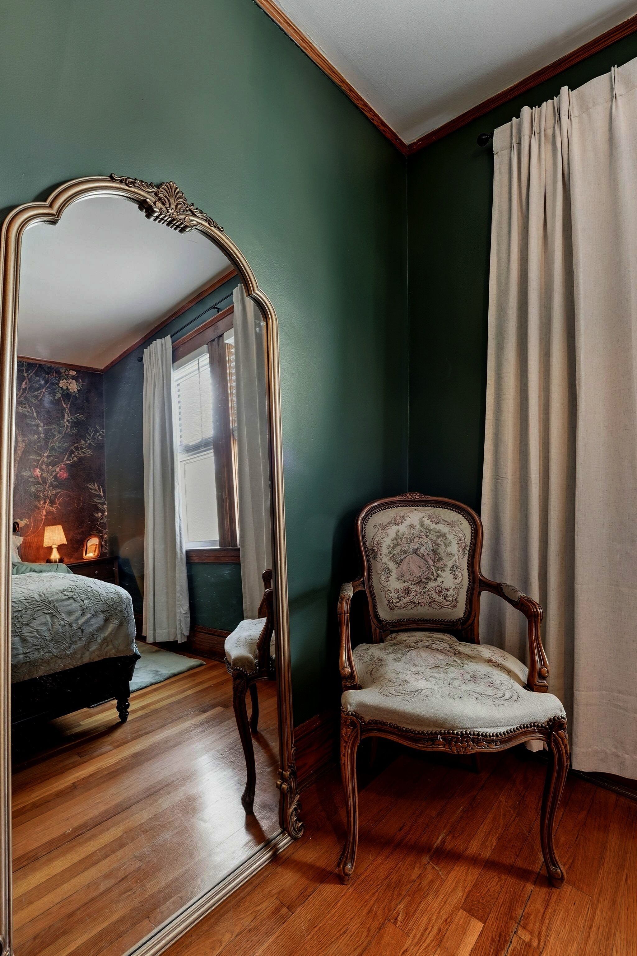 A room with a large mirror, a wooden floor, a chair, and a bed.