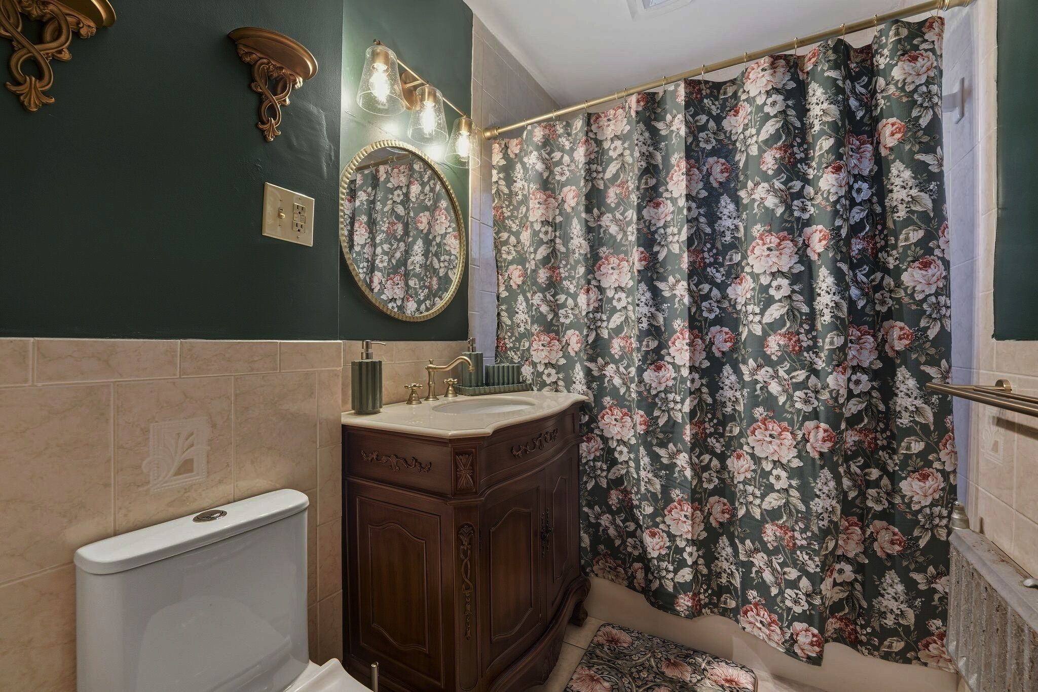 A bathroom with a dark green wall, a wooden vanity with a gold faucet, a white toilet, and a floral shower curtain.