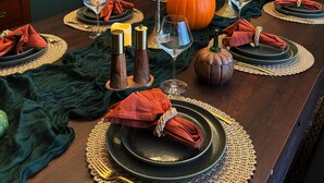 Mesa de comedor lista con platos, servilletas y velas, rodeada por una silla tapizada de verde y un aparador de madera.