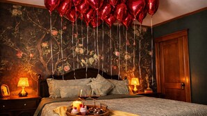 Un dormitorio con papel tapiz floral, una cama de madera, dos lámparas de noche, una mesita de noche con lámpara, una bandeja con velas y aperitivos, y globos con forma de corazón rojo.