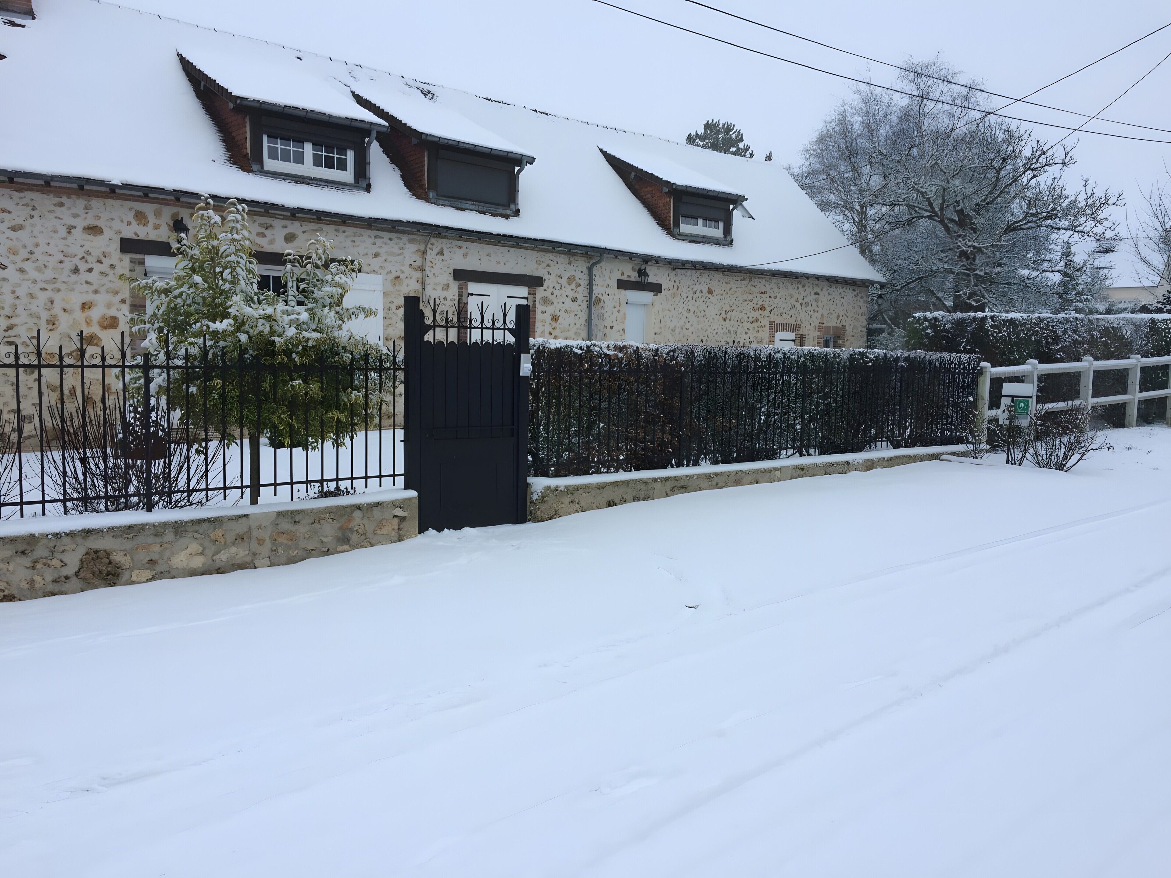 A snow-covered house with a stone facade, a black gate, and a wooden fence.