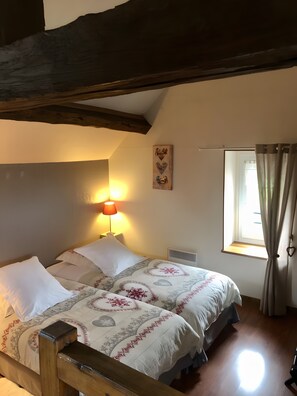 A bedroom with a wooden bed frame, two white pillows, a bedside table with a lamp, a window with curtains, and a wall-mounted artwork.