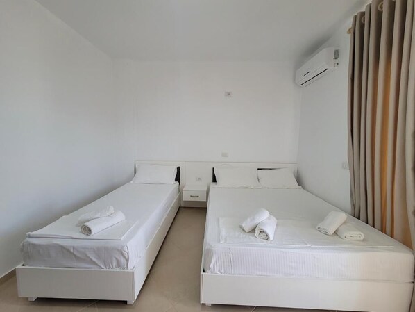 A room with two white beds, a small bedside table, a wall-mounted air conditioner, and a curtain on the right side. - Black Pearl Hotel (Ksamil)