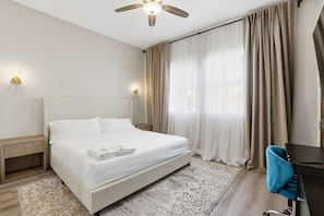 Une chambre d’hôtel avec un lit, un bureau, une chaise, un ventilateur de plafond et une fenêtre avec des rideaux.