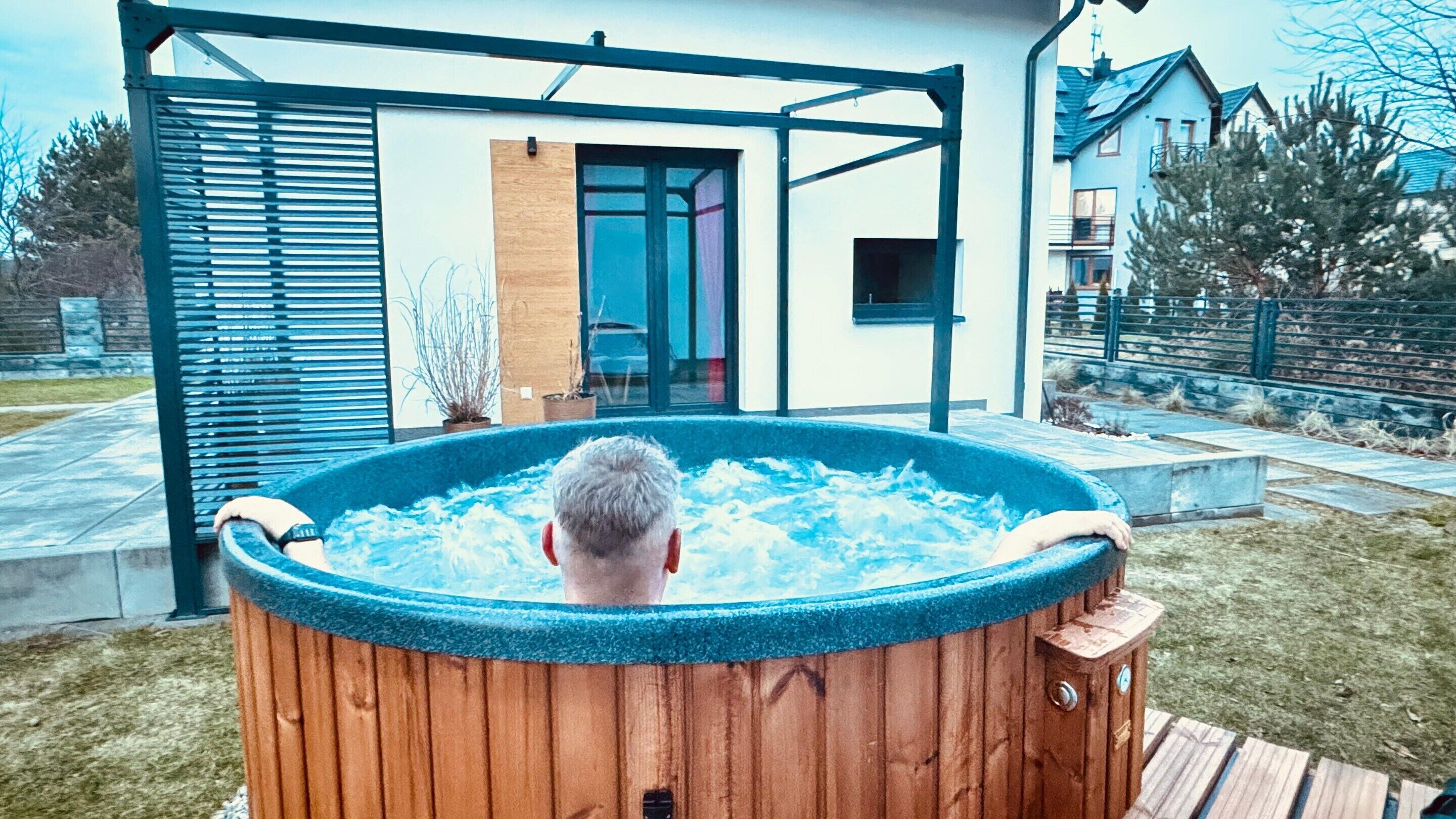 A wooden hot tub in a backyard with a modern house in the background.