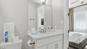 Un baño de hotel con inodoro, lavamanos, espejo y una cama visible a través de una puerta abierta.