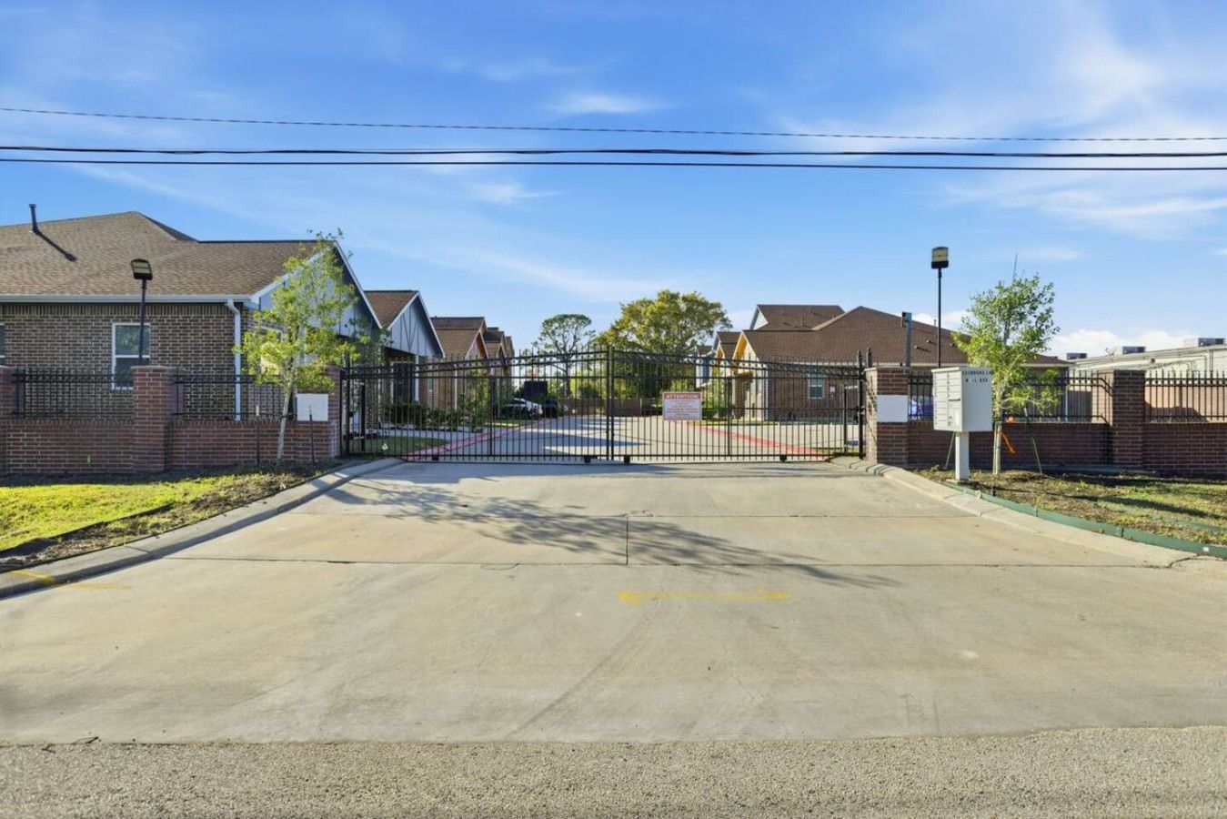 Une entrée fermée par un portail donnant accès à un quartier résidentiel avec des maisons en briques et un ciel dégagé.