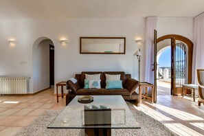 A living room with a brown sofa, a glass coffee table, and a view of the outdoors. - Villa rental in Moraira, MIMOSA (Moraira)