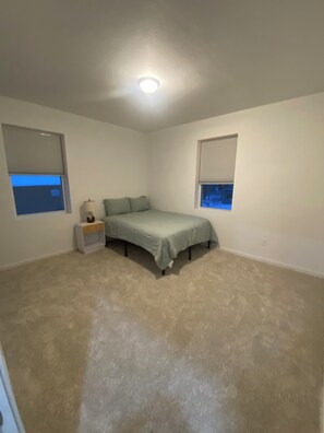 Une chambre à coucher comprenant un lit, deux tables de chevet, un luminaire au plafond et deux fenêtres équipées de stores.