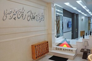 Un moderno vestíbulo de hotel con una mesa de recepción que dice "DAR AMIRA", una pared decorativa con caligrafía y un gran espejo que refleja el interior.