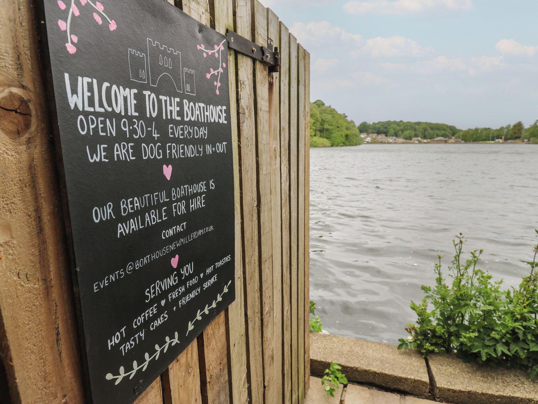 A sign welcoming visitors to a boathouse, open daily from 9:30 am to 4 pm, dog-friendly, and available for hire.