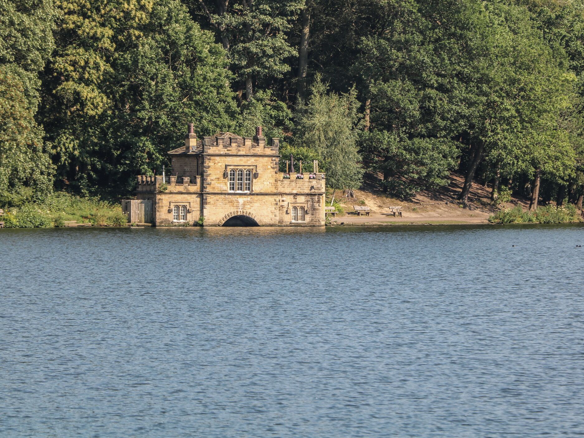 A stone building with a tower, located by a lake surrounded by trees.