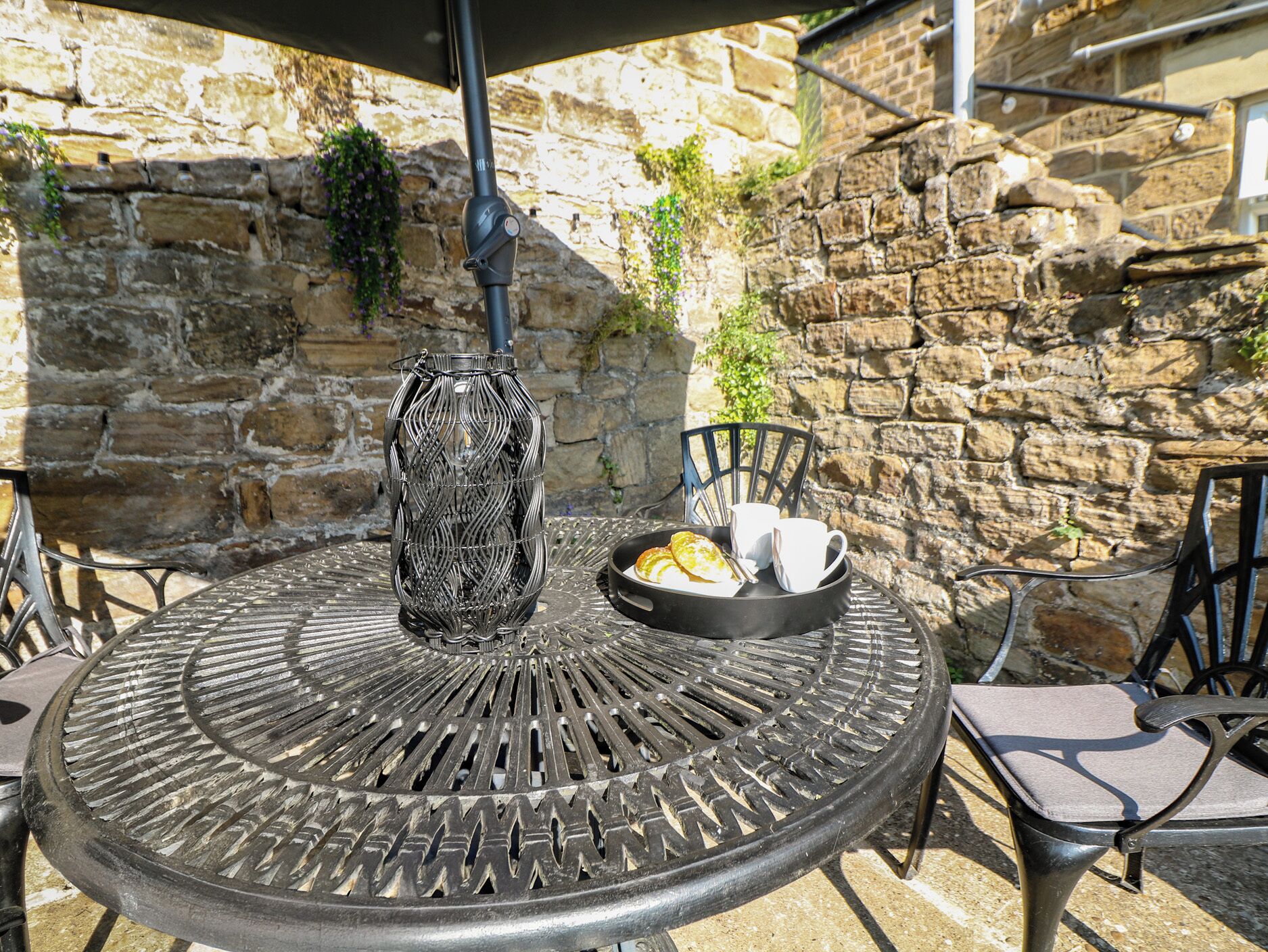 A round metal table with a decorative metal centerpiece, a black tray with a plate and cups, and a stone wall in the background.