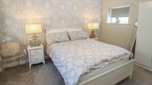 A bedroom with a bed, two bedside tables, lamps, a nightstand, a carpeted floor, and a window with a view. - Lake Side Cottage (Wakefield)